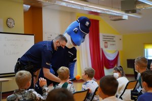 Policjant i Polfinek podczas rozmowy z dziećmi siedzącymi na krzesłach w sali .