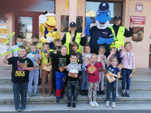 Zdjęcie grupowe uczniów, policjantów i maskotek. Wszyscy stoją na schodach wejściowych do budynku szkoły.