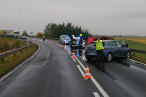 policjantka przeprowadza oględziny niebieskiego samochodu. W oddali stoją strażacy, radiowozy oraz policjant, który wstrzymuje ruch