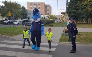 Polfinek idzie z dwojgiem dzieci przez przejście dla pieszych. obok stoi policjantka i obserwuje ich.