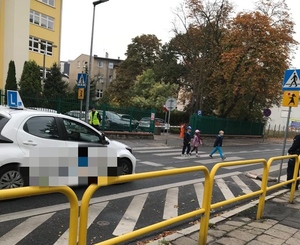 Dzieci przechodzą przez przejście dla pieszych pod okiem policjantów. Z lewej strony widoczny samochód osobowy ustępujący pierwszeństwa pieszym.