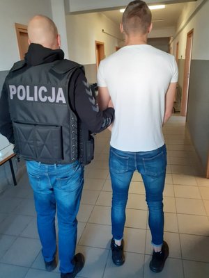 Policjant prowadzi zatrzymanego korytarzem komisariatu. Ujęcie z tyłu.