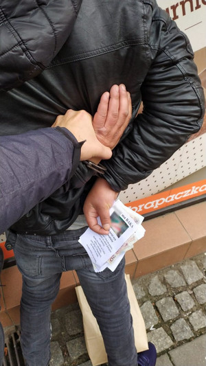 policjant trzyma zatrzymanego skutego kajdankami założonymi na ręce trzymane z tyłu. Mężczyzna trzyma w ręce ulotki oraz pieniądze