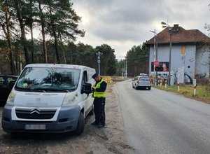 Policjant podczas kontroli drogowej samochodu dostawczego. W tle przejazd kolejowy.