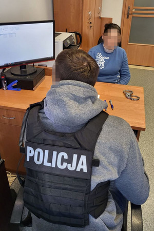 policjant siedzi na krześle przed biurkiem i patrzy w stojący obok monitor. Na przeciwko siedzi zatrzymana kobieta