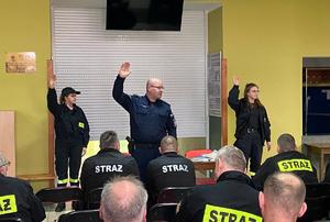 Sala szkoleniowa. Strażacy siedzą na krzesłach. Przed nimi stoi policjant oraz dwie kobiety strażaczki, którzy mają podniesione prawe ręce w górę.