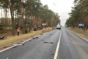 widok na drogę, gdzie leżą odłamki od rozbitych pojazdów. Na drodze pracują służby ratunkowe. Natomiast na poboczu obok drzew leży przewrócony na bok rozbity samochód