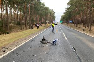 widok na drogę, gdzie leżą odłamki od rozbitych pojazdów. Na drodze pracują służby ratunkowe. Natomiast na poboczu obok drzew leży przewrócony na bok rozbity samochód