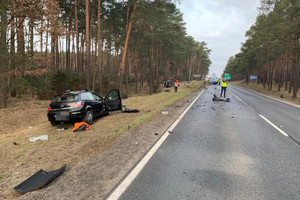 widok na drogę, gdzie leżą odłamki od rozbitych pojazdów. Na drodze pracują służby ratunkowe. Natomiast na poboczu obok drzew stoi rozbity opel, a dalej leży przewrócony na bok rozbity samochód