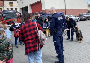 Policjanci , w tym jeden z policyjnym psem służbowym, stoją razem z grupą dzieci na terenie zewnętrznym jednostki pożarniczej.