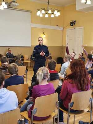 Policjantka stoi przed grupa siedzących uczniów i do nich mówi.