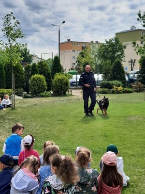 Policjant z psem służbowym stoi na terenie zielonym. Przed nim siedzą dzieci w półkolu.