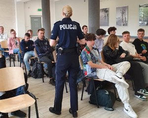 Policjantka stoi przy siedzących na krzesłach uczniów.