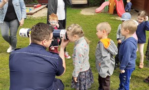 Policjant trzyma magiczną tubę, dziewczynka patrzy przez nią. Za nią stoją kolejne dzieci.