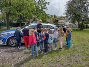 Policjantka rozmawia z dziećmi stojącymi przed nią w półkolu. Wszyscy stoją na tle radiowozu i zieleni.