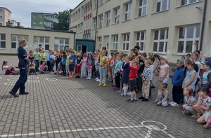 Policjantka rozmawia z dziećmi stojącymi w półkolu przed nią na terenie przed szkołą.