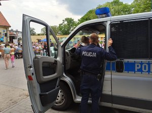 Policjant stoi przy otwartym radiowozie, w którym siedzi dziecko. W tle inny uczestnicy festynu.