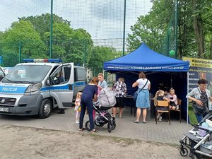 widok na namiot policyjny i stojący obok radiowóz. Przed nimi stoją osoby dorosłe i dzieci.