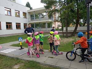 Dzieci na rowerach i hulajnogach jadą po torze. Przyglądają się temu policjanci.