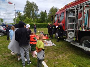 Dzieci oglądają sprzęt strażacki rozłożony na trawie przed wozem bojowym.