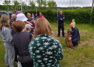 Policjant z psem służbowym kuca przed dziećmi stojącymi w półokręgu. Przygląda się temu drugi policjant.