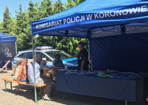 Osoby siedzą przy stoliku pod namiotem policyjnym, pod którym stoi policjantka.