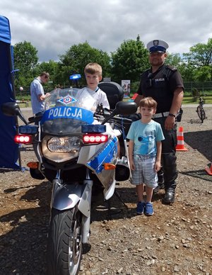 Policjant ruchu drogowego stoi przy służbowym motocyklu, obok niego stoi chłopiec. Na motocyklu siedzi drugi chłopiec.