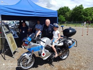 Przed policyjnym namiotem profilaktycznym stoi motocykl policyjny. Na nim siedzi dziecko. Przy motocyklu stoi policjant.