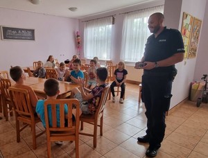 Policjant rozmawia z dziećmi siedzącymi przy stolikach.