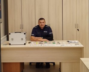 Policjant siedzi przy biurku, na którym jest walizka i rozłożone atrapy substancji narkotycznych.