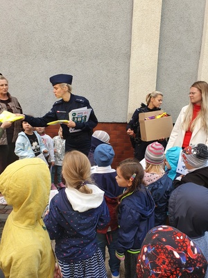 Policjantka rozdaje kamizelki odblaskowe stojącym przed nią  dzieciom.