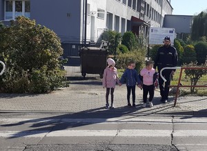 Policjant wraz z dziećmi stoją przy przejściu dla pieszych. W tle budynek.