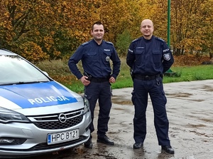 Policjanci stojący obok radiowozu w terenie zielonym.