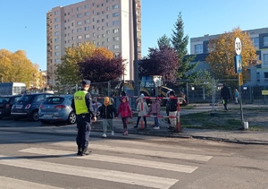 Dzieci ćwiczą przechodzenie przez przejście dla pieszych w rejonie szkoły pod okiem policjanta.