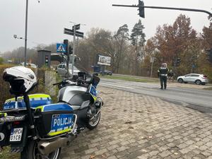 Policjant kieruje ruchem w rejonie przejścia dla pieszych. Na pierwszym planie policyjny motocykl.