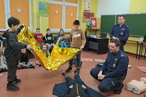 dwójka chłopców trzyma folie termiczną, na ziemi leży dziecko. Obok klęczy policjant, który tłumaczy udzielanie pomocy