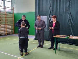 Policjant wręcza nagrody chłopcu. Obok stoją dwie dorosłe osoby.