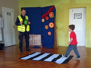 chłopiec przechodzi przez matę imitującą przejście dla pieszych, przed nim stoi policjant trzymając planszę z zielonym światłem drogowym