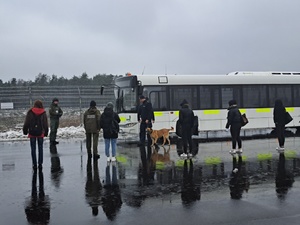 Policjant z psem oraz inne osoby na terenie otwartym lotniska, przed nimi stoi autobus.