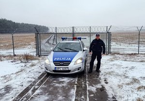 policjant stoi przy radiowozie zaparkowanym przy ogrodzeniu lotniska