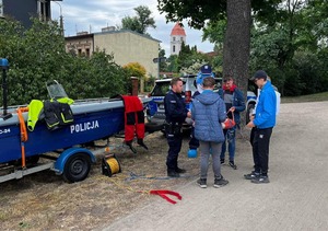 policjant rozmawia z osobami, w tle radiowóz i policyjna łódź