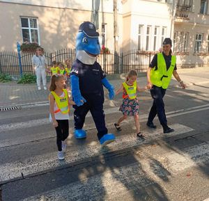 Polfinek i policjantka przechodzą z dziećmi przez przejście dla pieszych