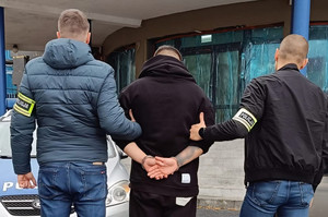 Policjanci prowadzą zatrzymanego mającego założone kajdanki na ręce trzymane z tyłu.